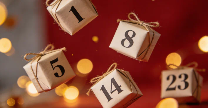 Adventskalender-Zahlen auf kleinen Geschenkboxen vor weihnachtlichem Lichterhintergrund – Symbolbild für einen erotischen Adventskalender 2026.