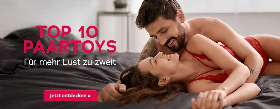 Ein Paar, welches sich anlächelt, liegt auf einem Bett mit dunkler Decke. Die Frau trägt rote Spitzenunterwäsche, der Mann liegt halb über ihr und umarmt sie. Großflächig steht der Text „Top 10 Paartoys – Für mehr Lust zu zweit“. Unten roter Button mit weißer Schrift „Jetzt entdecken ».