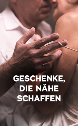 Das Bild zeigt eine Werbeanzeige, wo eine Frau und ein Mann dicht aneinander stehen. Der Mann trägt ein weißes Hemd und die Frau ein helles Oberteil mit dünnen Trägern. Die Köpfe sind dicht beieinander und der Mann macht mit seiner Hand den dünnen Träger der Frau an ihrer Schulter runter. Plakativ steht in großer, weißer Schrift "Geschenke, die Nähe schaffen". Unten befindet sich ein rotfarbener Button mit der weißen Aufschrift „Zeig’s mir“. Der Hintergrund ist dunkel gehalten.